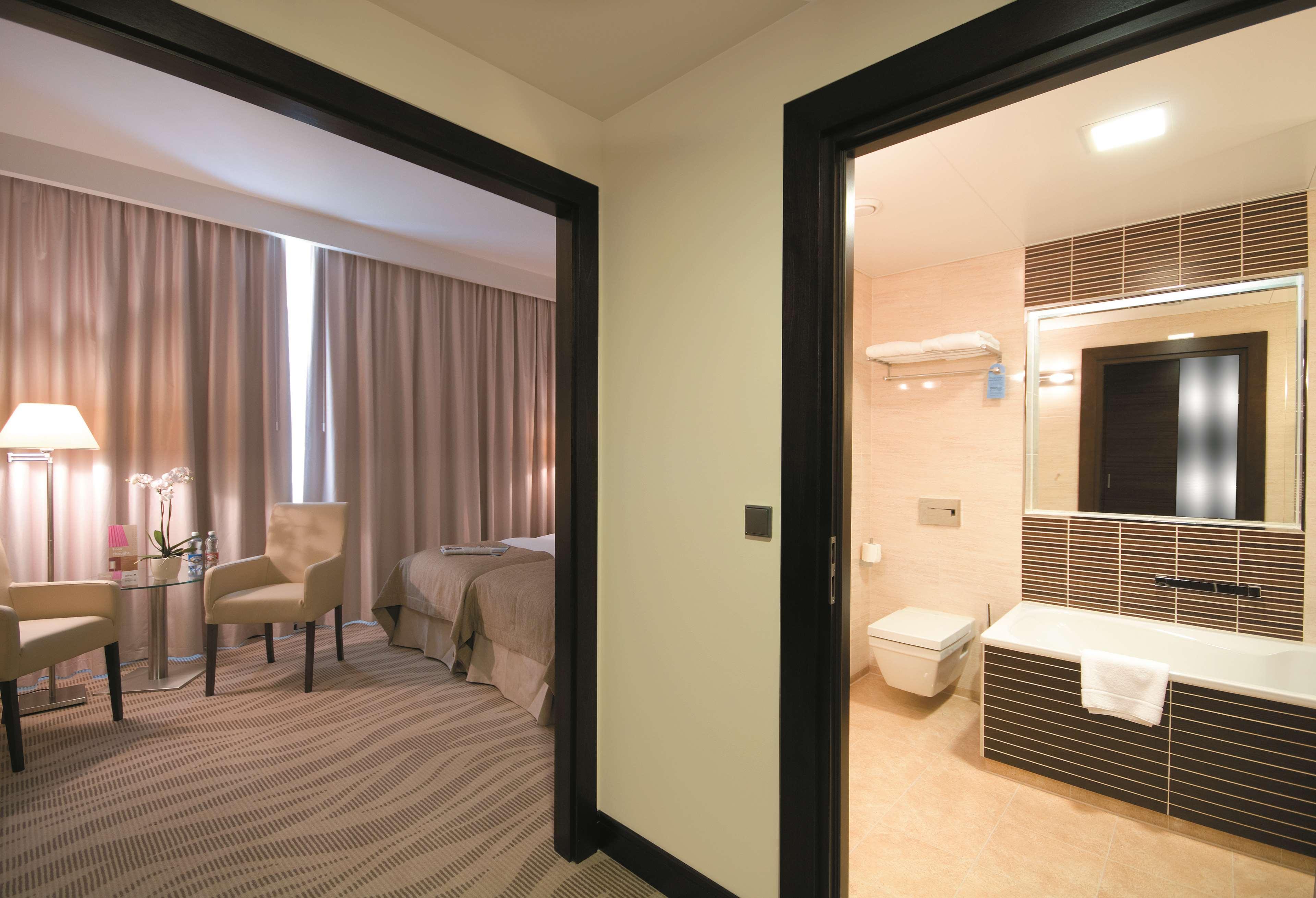 格但斯克，雷迪森布鲁酒店 外观 照片 The photo shows a view of a room with two doors leading to different areas. On the left side, there's a bedroom featuring a bed with a light gray coverlet, complemented by curtains and a small seating area with a chair and a table. On the right side,
