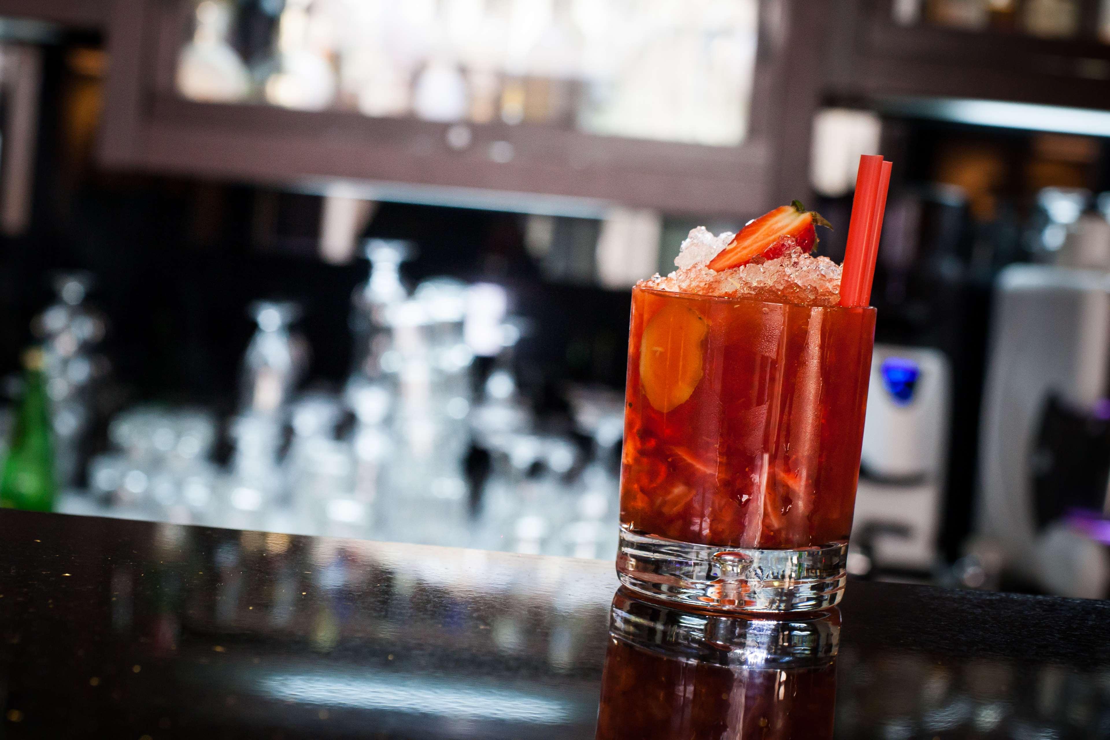 格但斯克，雷迪森布鲁酒店 外观 照片 The photo shows a glass of a colorful cocktail placed on a dark surface, likely a bar countertop. The drink is garnished with slices of fruit, possibly strawberries, and is filled with ice. In the background, there are blurred outlines of other glass