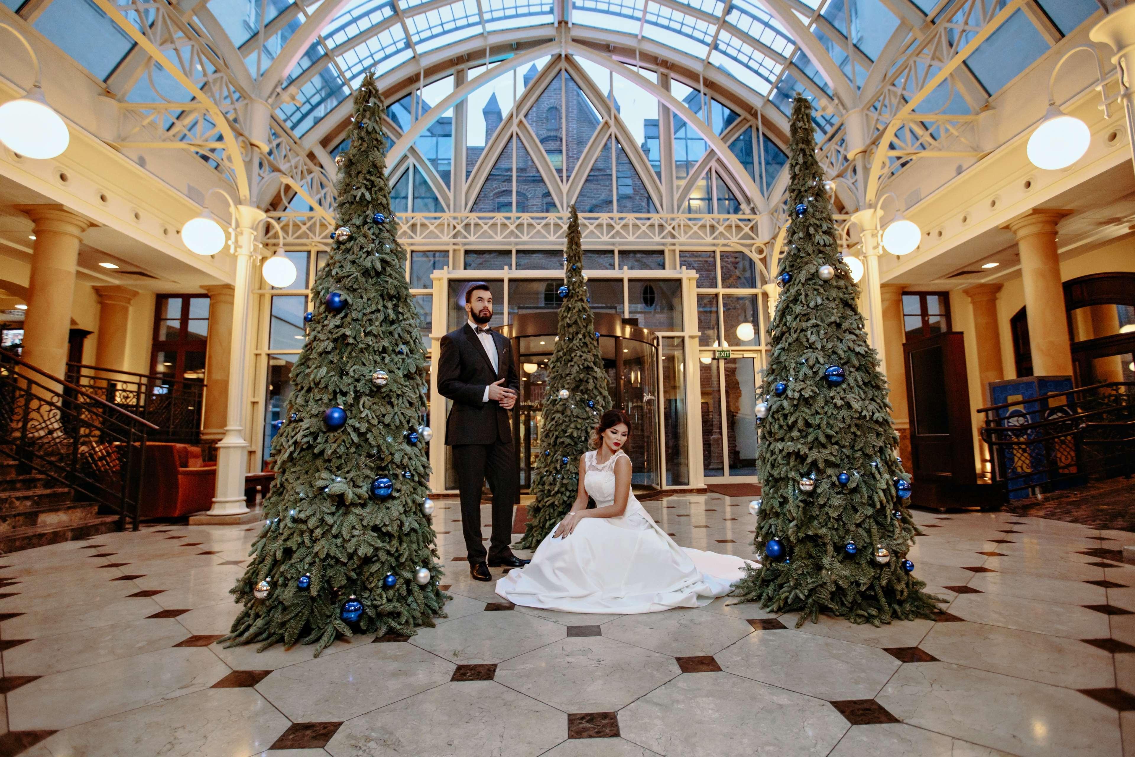 格但斯克，雷迪森布鲁酒店 外观 照片 The photo shows a groom in a tuxedo standing next to a bride in a white wedding dress. They are positioned in a beautifully decorated indoor space, which features large glass windows and elegant architectural details. Surrounding them are three green