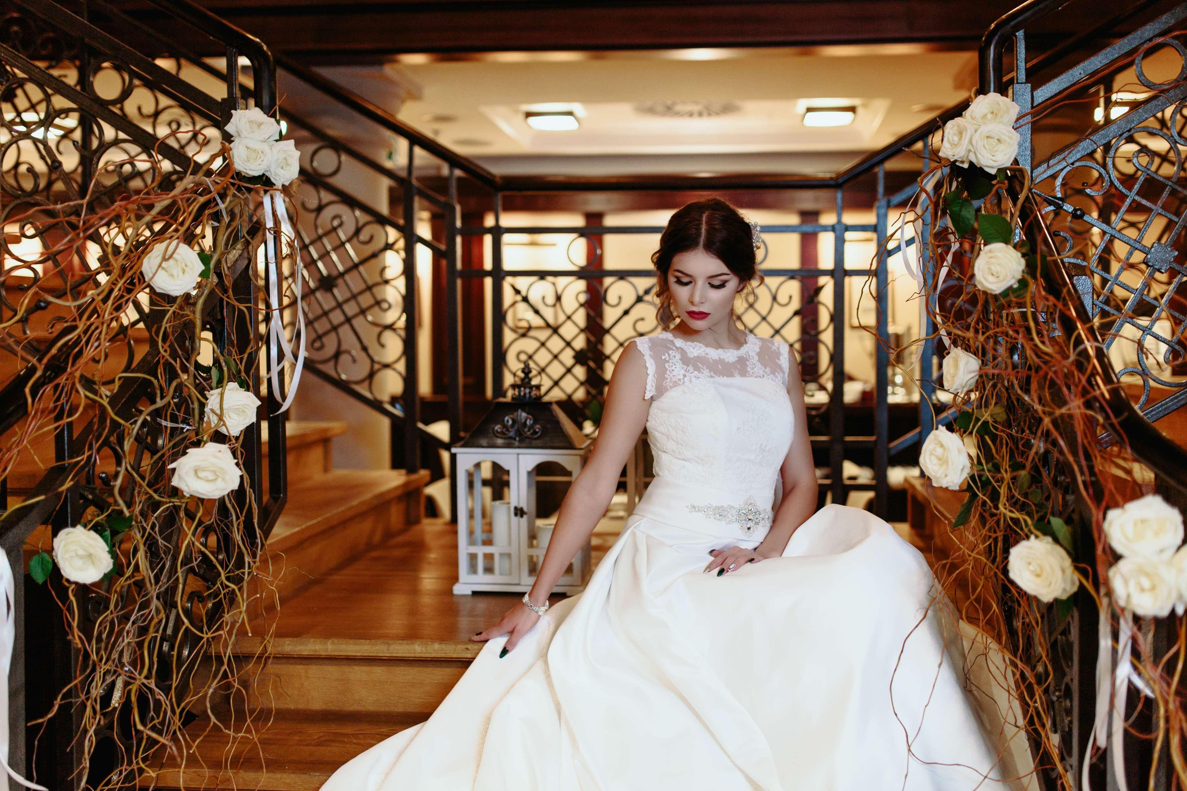 格但斯克，雷迪森布鲁酒店 外观 照片 The image shows a bride seated on a staircase in an elegant indoor setting. She is wearing a beautiful white wedding gown with intricate details, including a sheer neckline. The staircase is adorned with decorative elements, such as white roses and f