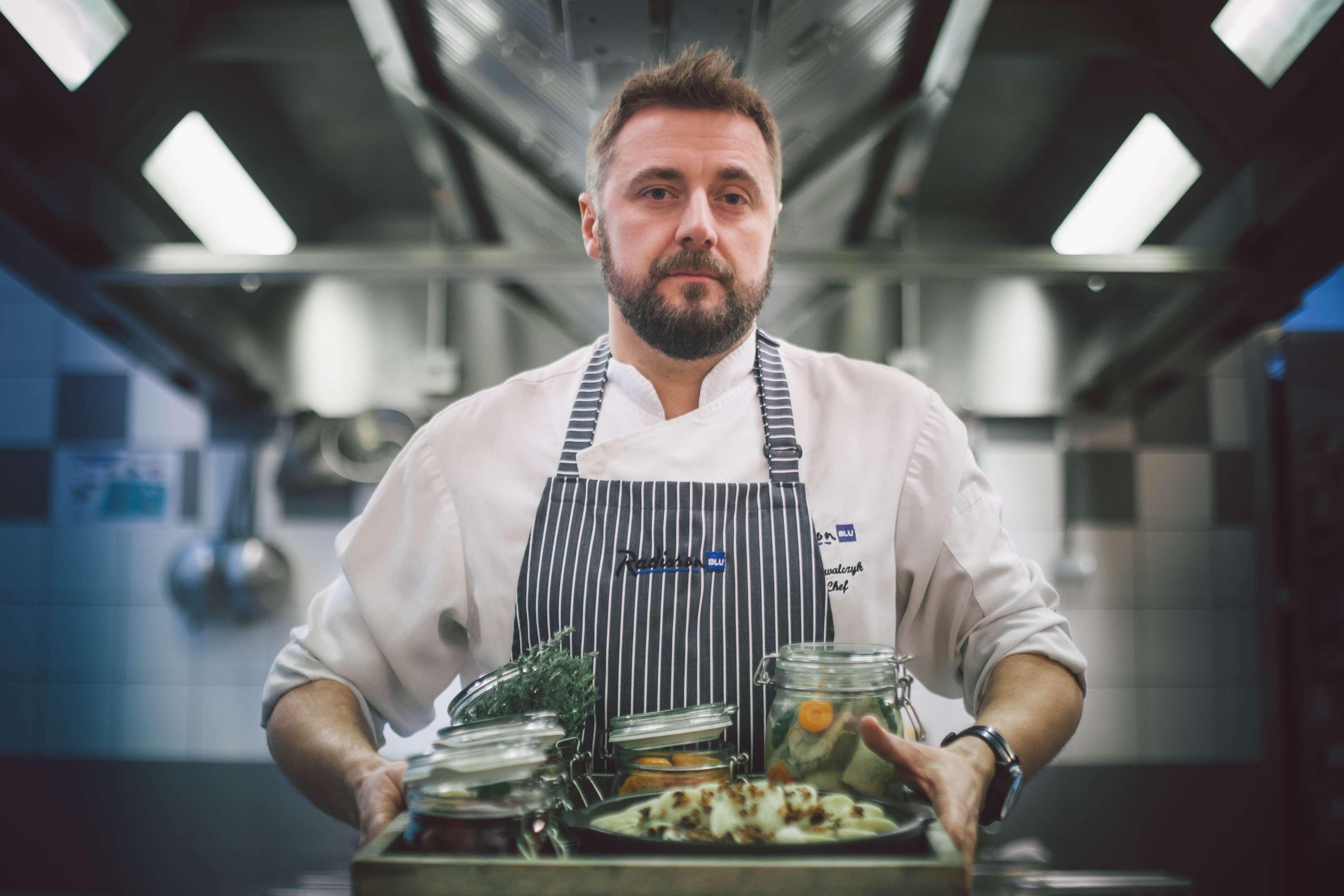 格但斯克，雷迪森布鲁酒店 外观 照片 The photo depicts a chef in a professional kitchen setting. He is wearing a white chef's coat with a striped apron and has a focused expression. He is holding a wooden tray filled with various jars and containers that likely contain ingredients for c