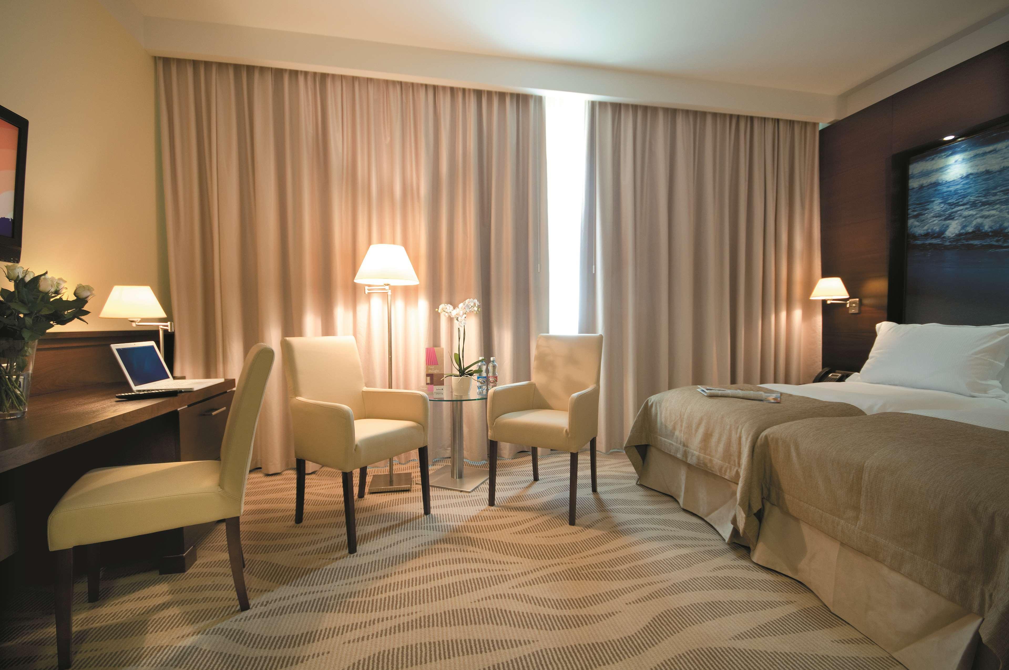 格但斯克，雷迪森布鲁酒店 外观 照片 The photo shows a hotel room that features a queen-sized bed with neutral-colored bedding. There are two chairs and a small glass table in front of the bed. A desk is situated to one side, likely for work or personal use. The room has two lamps provi