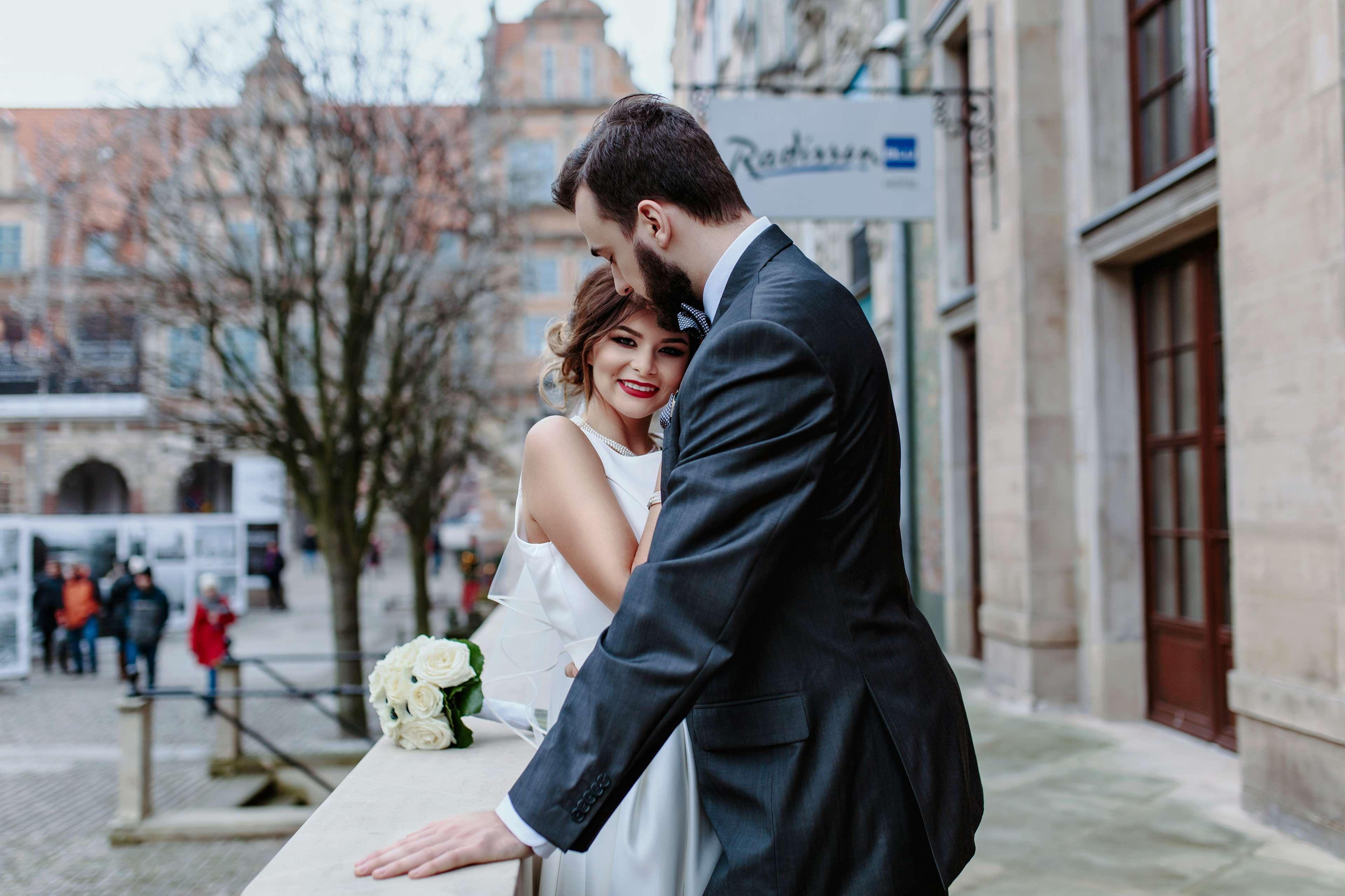 格但斯克，雷迪森布鲁酒店 外观 照片 The photo shows a couple embracing in an outdoor setting. The woman, dressed in a white wedding gown, is smiling and appears to be enjoying the moment. The man, wearing a dark suit, leans in toward her, suggesting a romantic atmosphere. In the backgr