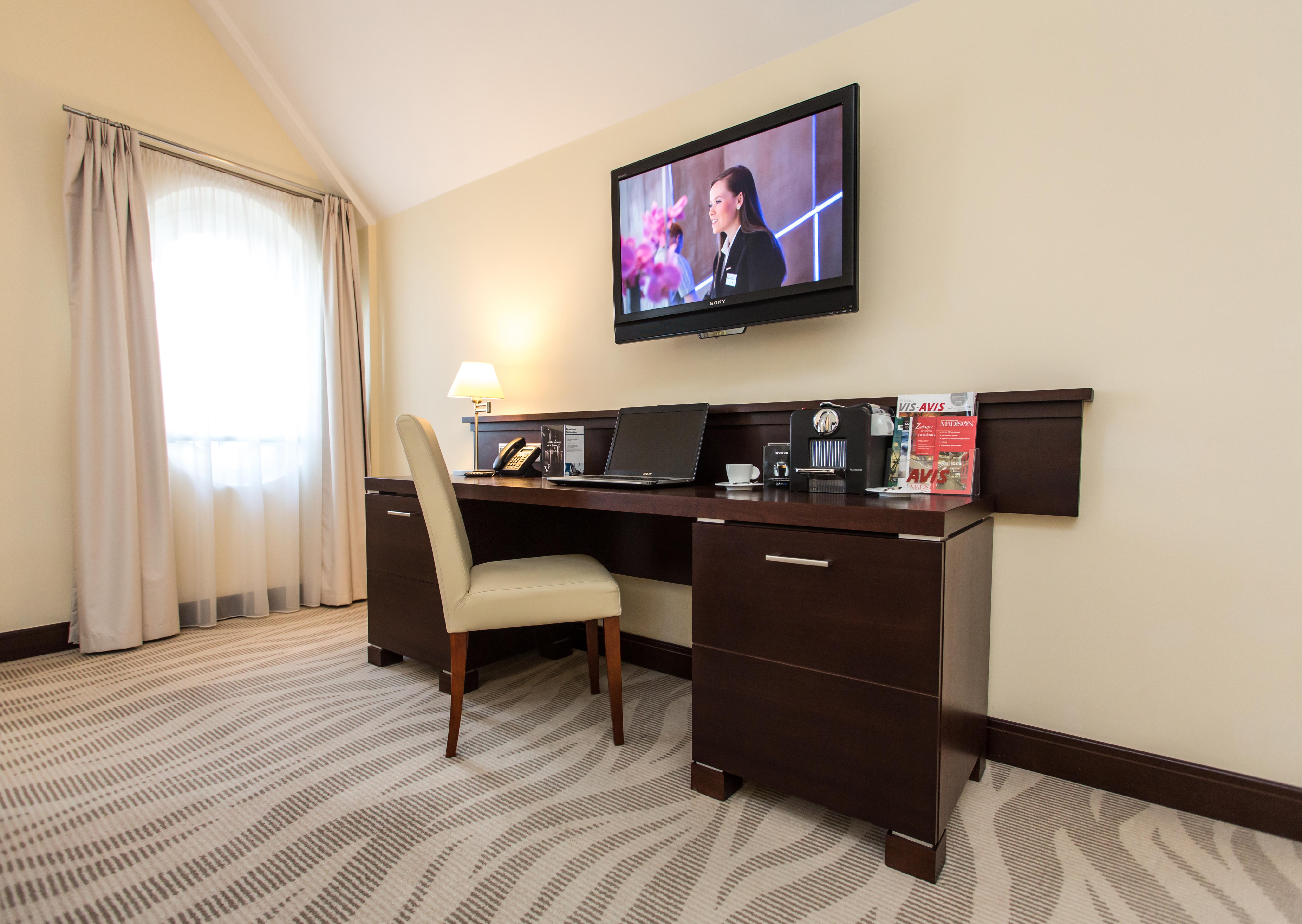 格但斯克，雷迪森布鲁酒店 外观 照片 The photo depicts a corner of a room featuring a stylish desk with a chair and a television mounted on the wall above. The desk has various items on it, possibly including a telephone, clock, and some decorative elements or stationery. There is a lam