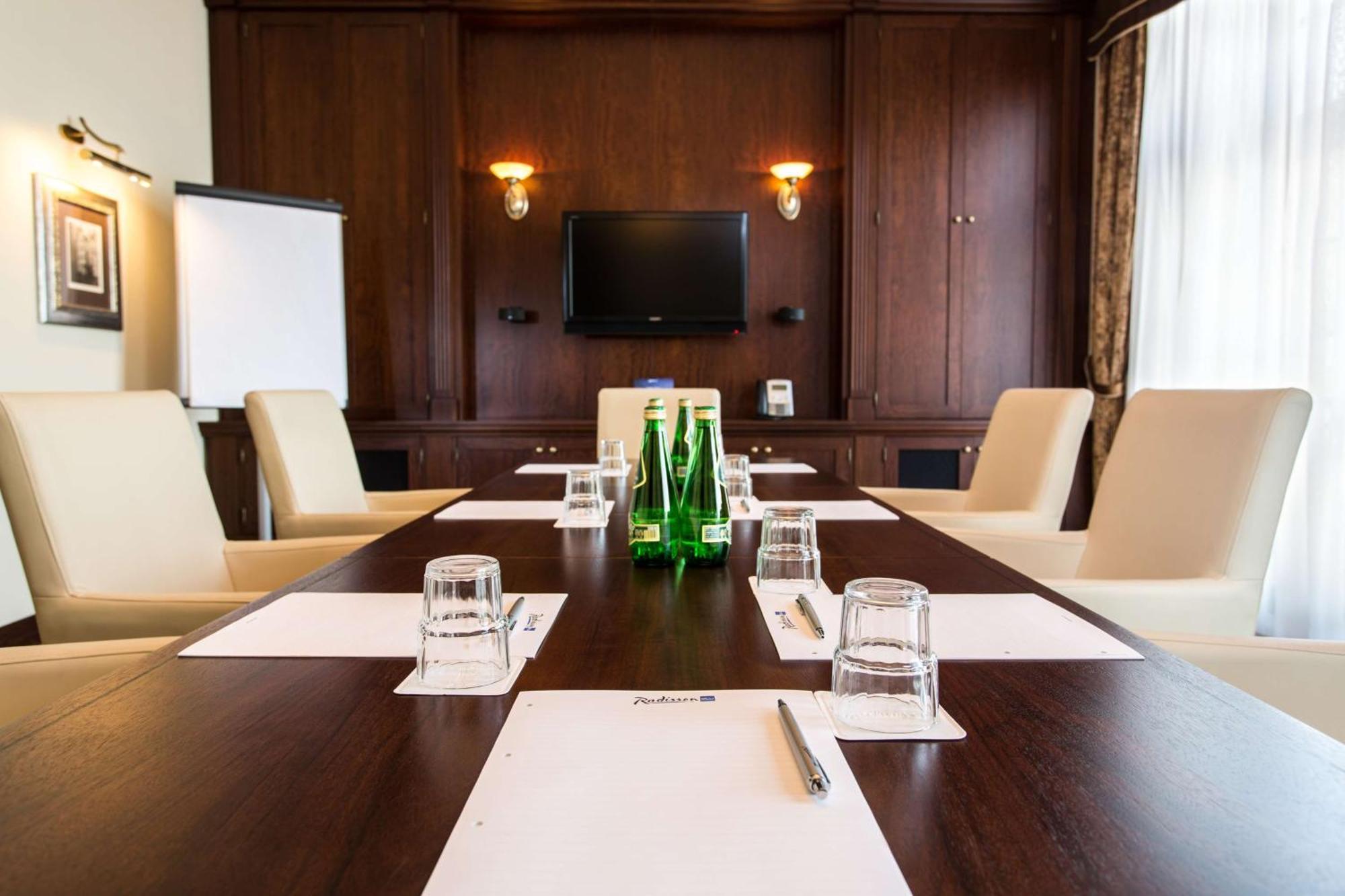 格但斯克，雷迪森布鲁酒店 外观 照片 The photo shows a conference room setup. There's a large wooden table with several cream-colored chairs around it. On the table, there are sheets of paper, water glasses, and small bottles of water. A flat-screen television is mounted on the wall at 