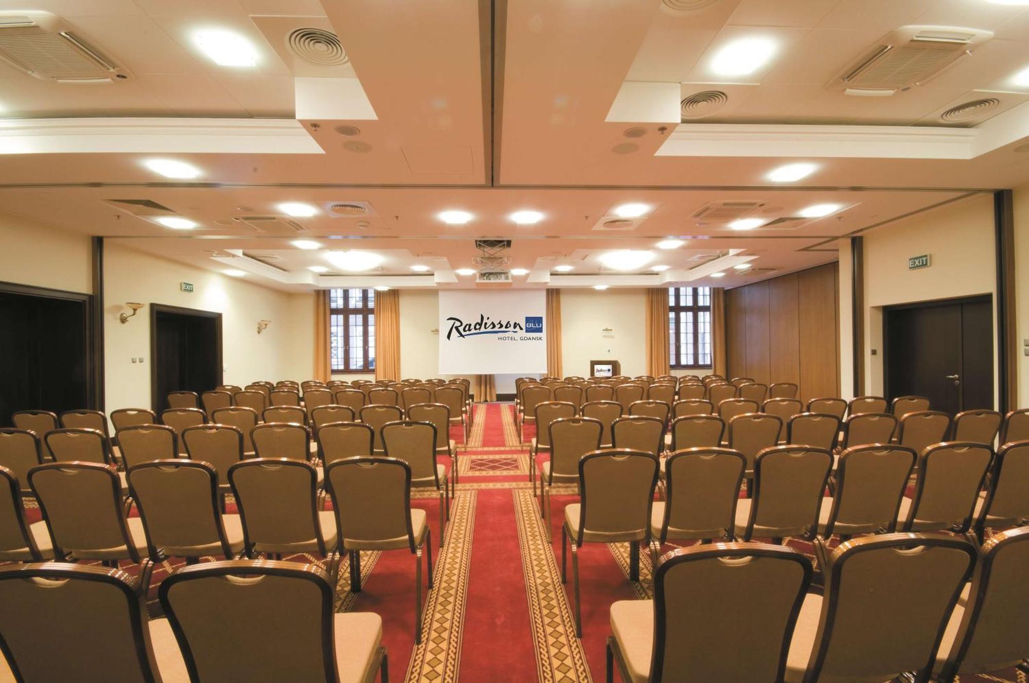 格但斯克，雷迪森布鲁酒店 外观 照片 The photo shows the interior of a conference room or event space. It features rows of brown chairs arranged neatly, facing towards the front where a stage or presentation area is set up. There is a carpet running down the center of the room, and the 