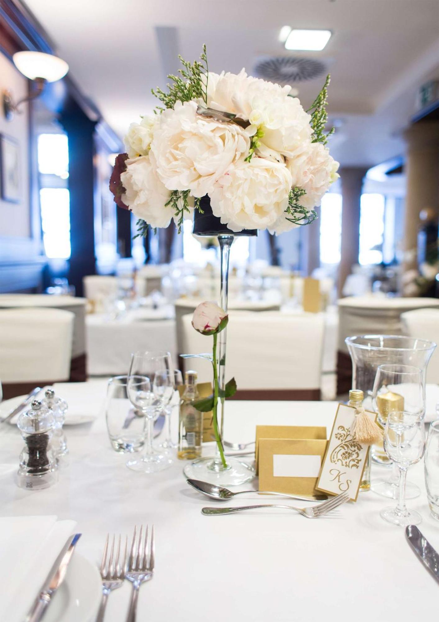 格但斯克，雷迪森布鲁酒店 外观 照片 The photo depicts a beautifully arranged table setting, likely for a formal event or celebration. At the center of the table is a tall floral arrangement featuring soft pink peonies and greenery, elegantly placed in a clear vase. Surrounding the cent