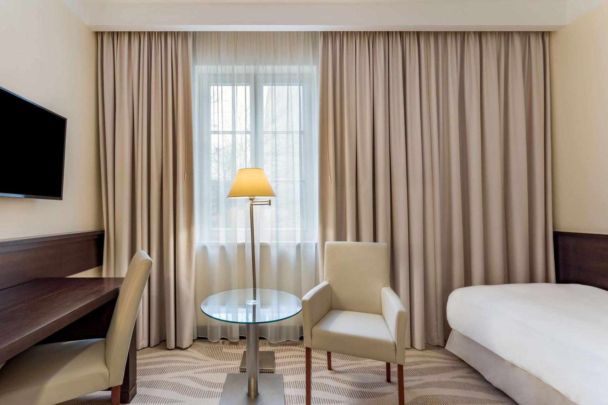格但斯克，雷迪森布鲁酒店 外观 照片 The photo shows a hotel room that features a light and neutral color scheme. There is a window covered with sheer curtains, allowing natural light to enter the space. To the right, there is a bed with a white bedspread. 

In the center, there is a sm