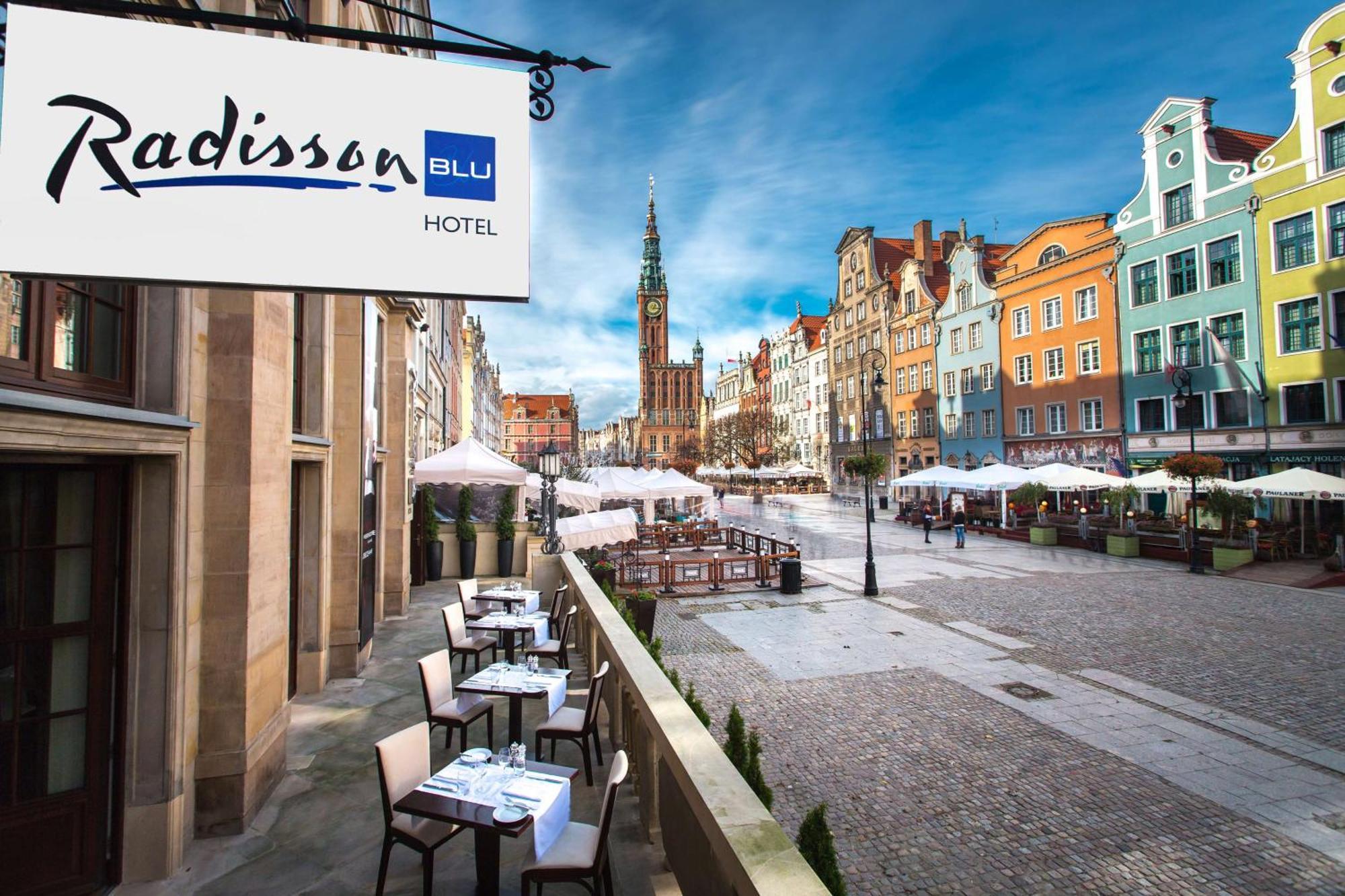 格但斯克，雷迪森布鲁酒店 外观 照片 The photo shows a view of the Radisson Blu Hotel, which is situated in a vibrant, picturesque area. The image features an outdoor seating arrangement with tables and chairs, likely part of a restaurant or café. In the background, colorful historic bu