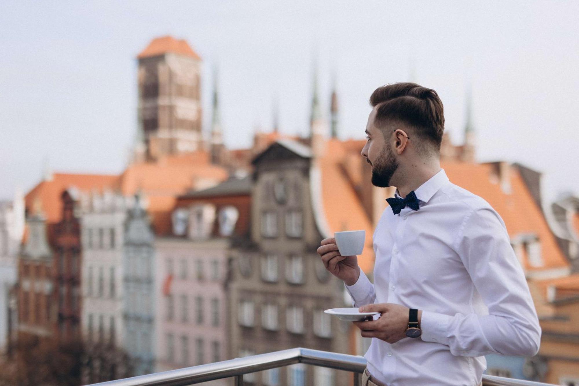 格但斯克，雷迪森布鲁酒店 外观 照片 The photo features a man standing on a balcony, holding a cup of coffee or tea in one hand while resting a saucer in the other. He is dressed in a white shirt with a bow tie. The background reveals a picturesque view of an old town, showcasing buildi